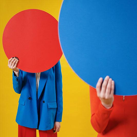 Kaksi värikkäisiin vaatteisiin pukeunutta ihmistä pitää kasvojen edessä punaista tai sinistä ympyrää