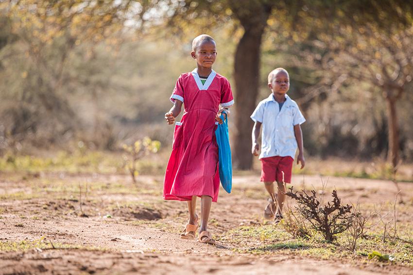 Tyttö ja poika koulupuvuissa kävelemässä kuivalla ja aurinkoisella seudulla.